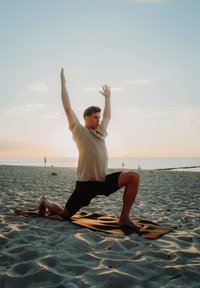 Osoba u bežoj košulji i crnim kratkim hlačama izvodi joga položaj na tamnom prostircu na plaži. U pozadini se vide mekani, šareni pijesak i zalazak sunca.