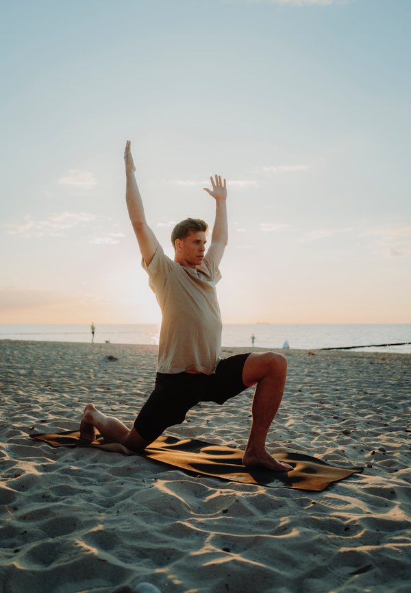 Una persona con una camiseta beige y pantalones cortos negros realiza una postura de yoga sobre una esterilla oscura en la playa. Se puede ver arena suave y con patrones y un fondo de atardecer.