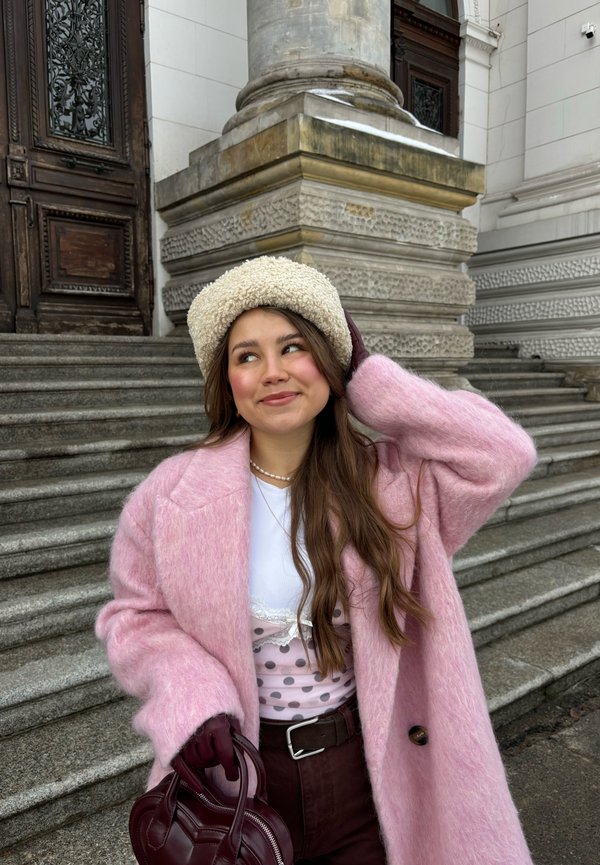 Jeune femme en manteau rose et chapeau crème debout sur des marches en pierre près d'une grande porte en bois ornée et d'une colonne. Elle tient un sac bordeaux.
