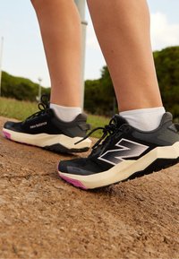 Des sneakers New Balance noirs présentent une tige en mesh avec une semelle rose et noire, un branding bien visible et un motif texturé sur le matériau.