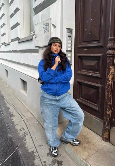 Joven con una chaqueta azul, jeans anchos y un gorro de lana, de pie con un pie en un escalón cerca de una puerta marrón y una pared blanca con grafitis.