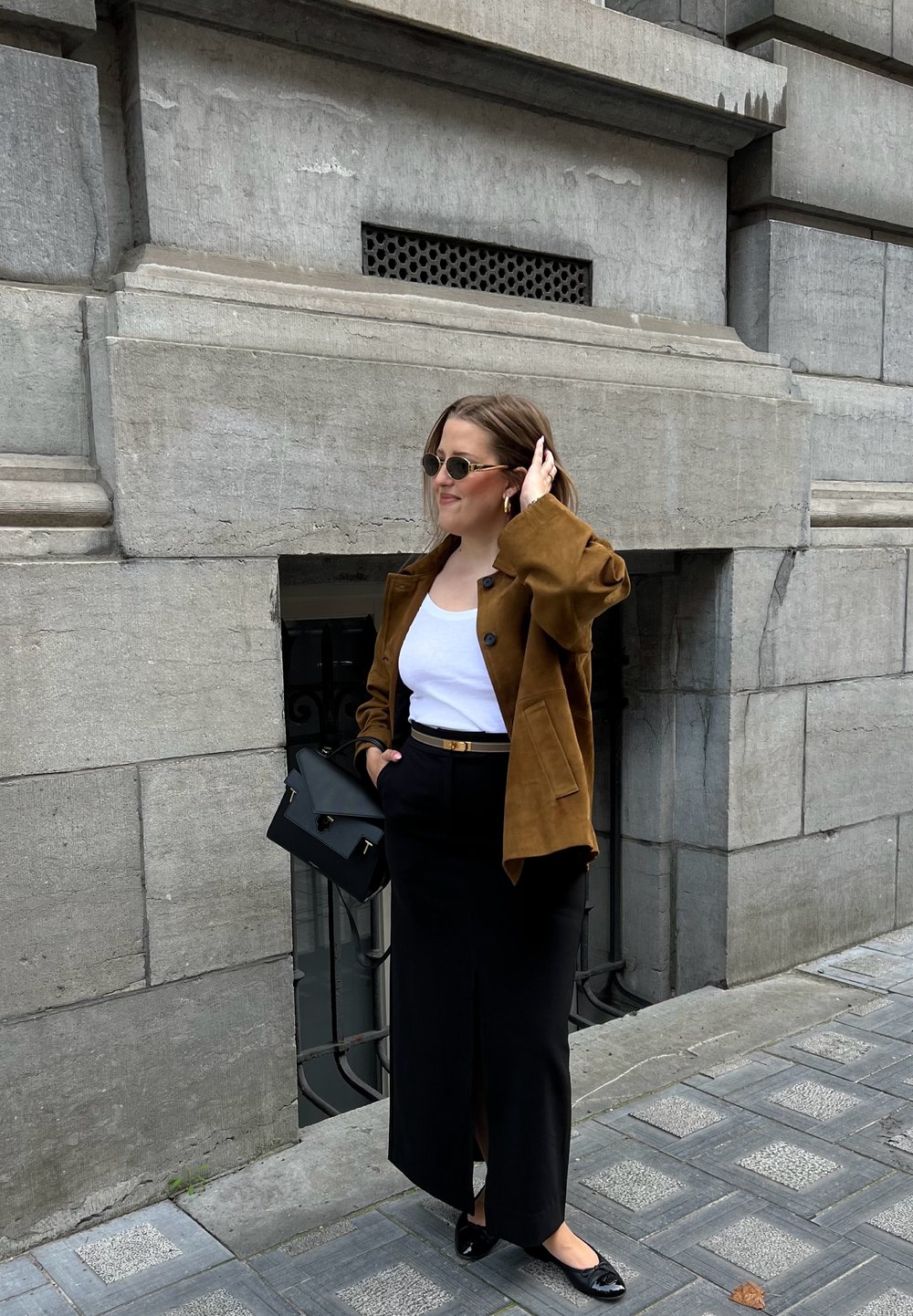 Pruunest nahast jope, valgest tipust, mustast kõrge vöökoha seelikust, millel on eeslõhik, mustad korterjalatsid ja must käekott kullatud detailidega halli seina taustal.