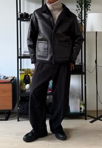 Veste en cuir noir avec un col en fourrure retournée, poches avant et fermeture par boutons, associée à un pantalon noir ample et des chaussures noires.