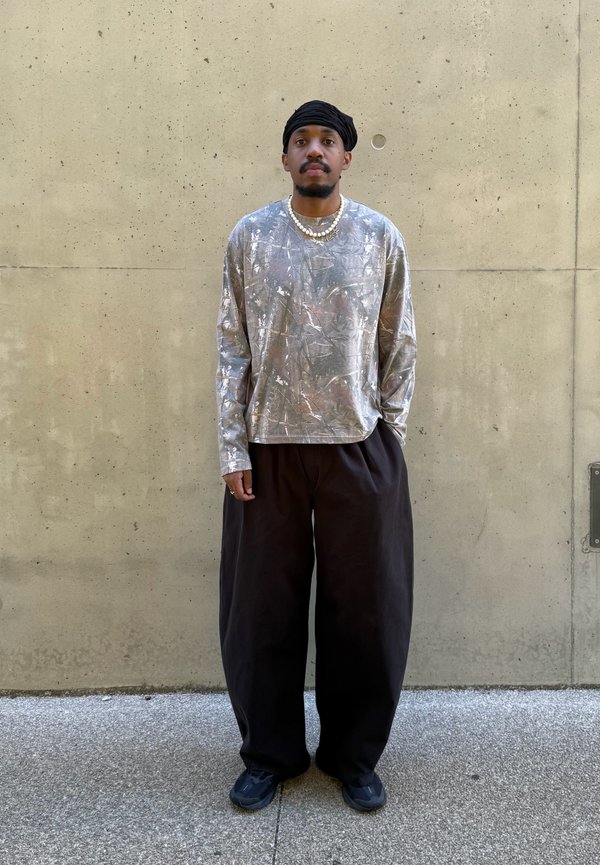 Homme portant un turban noir, une chemise à manches longues argentée avec des motifs, un pantalon large noir, des chaussures noires et des colliers de perles, debout contre un mur beige.