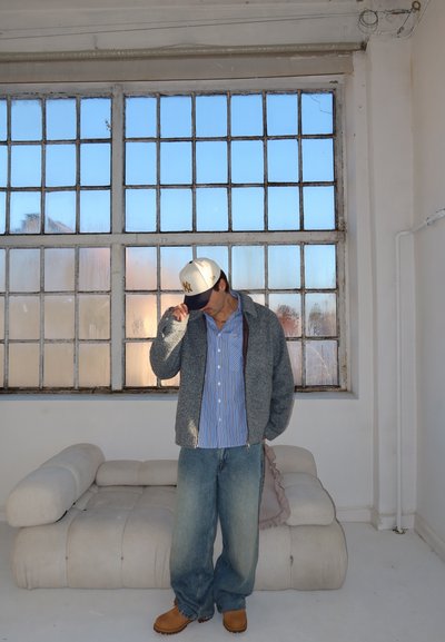 Hombre con atuendo casual que lleva chaqueta gris, camisa azul a rayas, vaqueros holgados, botas marrones y gorra blanca, de pie dentro de una habitación junto a una gran ventana y un sofá beige.