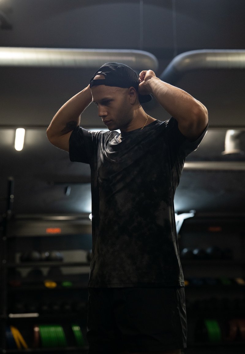 T-shirt atletica nera con un design testurizzato e a motivi. Chi lo indossa regola un cappellino nero contro uno sfondo sfocato di una palestra.