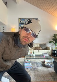 Polstret brun vest over en mørk høyhalset genser, beige strikkelue med svart mønster og hvite solbriller, satt i et moderne oppholdsrom.