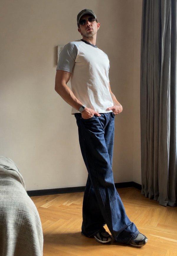 Homme portant des lunettes de soleil, une casquette, un t-shirt blanc, un jean bleu ample et des baskets, debout à l'intérieur près de rideaux gris et d'un sol en bois.