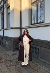 Brun lang kåpe over en kremfarget genser og matchende bukser. Brune, lodne tøfler, holder en teksturert grå håndveske. Urbant miljø med steinlagt fortau.