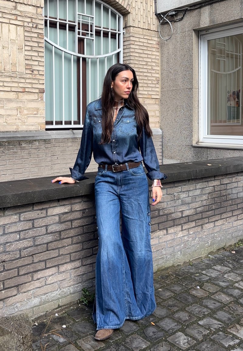 Femme aux longs cheveux foncés portant une chemise en denim et un jean à jambes larges, appuyée contre un mur de briques dans un cadre urbain avec des fenêtres derrière.