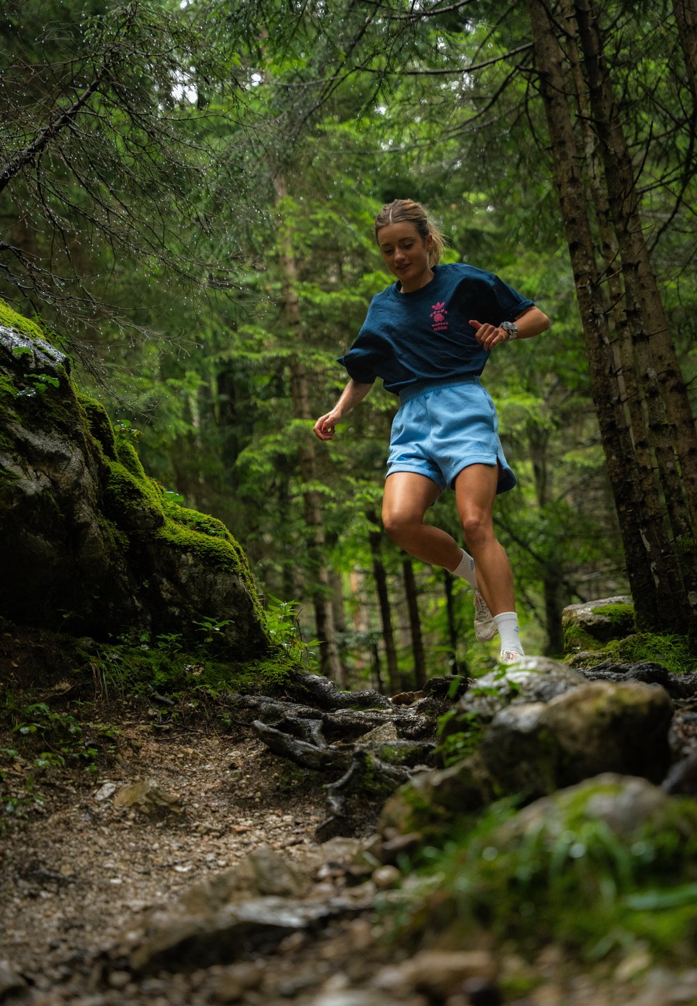 Blå shorts og en mørk t-shirt med et pink grafisk design, løbende på en stenfyldt, jordsti i en grøn skov, omgivet af træer og mos.