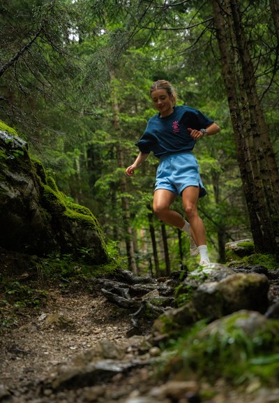 Pantalones cortos azules y una camiseta oscura con un gráfico rosa, corriendo por un sendero de tierra rocosa en un bosque verde, rodeado de árboles y musgo.