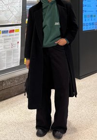 Long manteau noir par-dessus un sweat à capuche vert, associé à un pantalon noir à jambes larges et des chaussures noires. Textures unies, coupe décontractée.