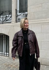 Veste en cuir bordeaux avec une fermeture éclair et des boutons-pression, portée par-dessus un haut noir, associée à un pantalon noir taille haute et un sac à main texturé.