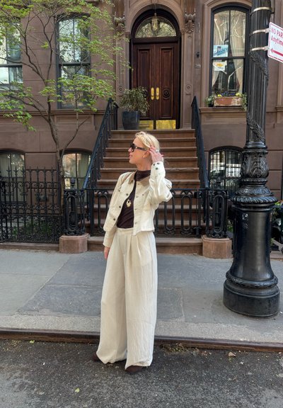 Mujer rubia con chaqueta beige y pantalones anchos está de pie en la acera de la ciudad frente a una casa de piedra marrón con escaleras y barandillas de hierro negro.