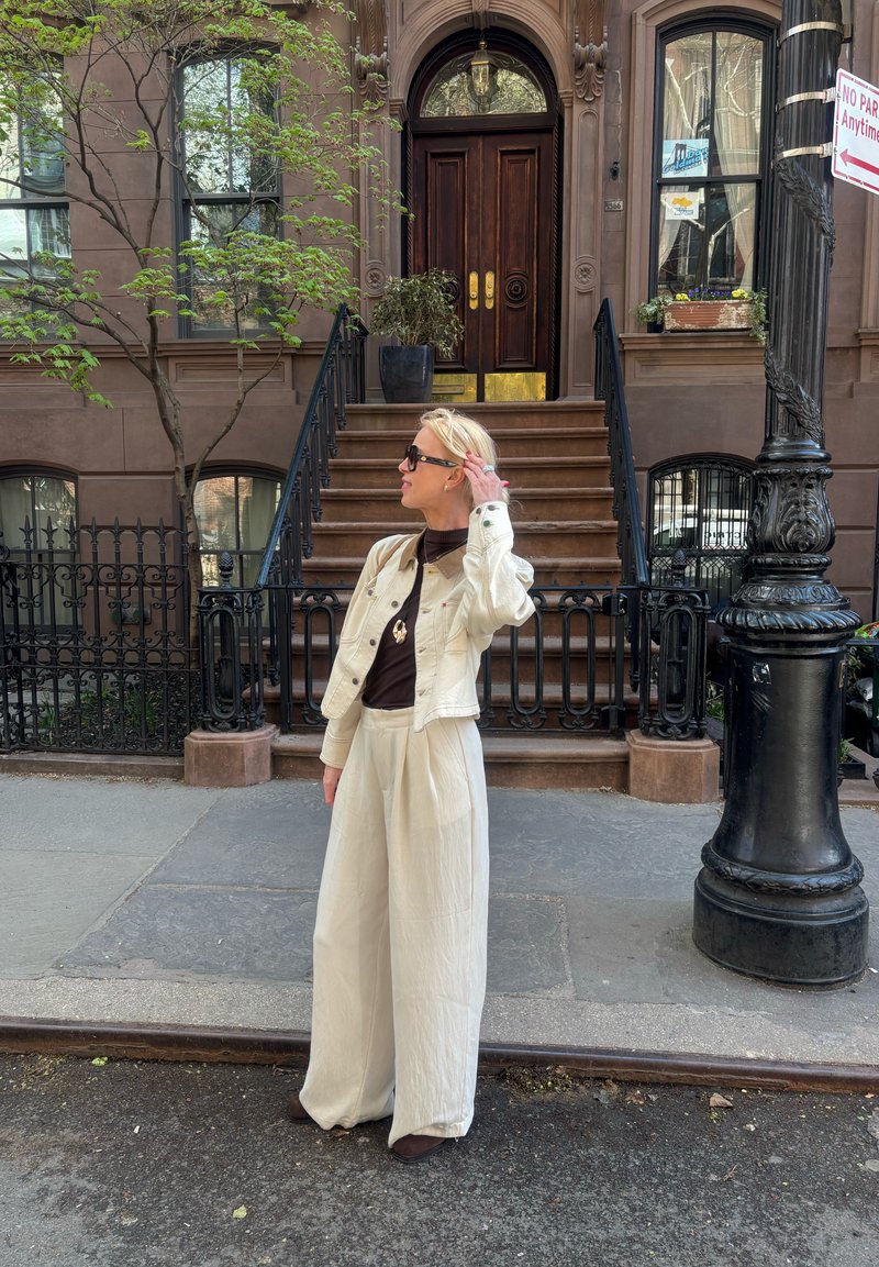 Femme blonde en veste beige et pantalon large debout sur le trottoir de la ville devant une maison en pierre brune avec des escaliers et des rampes en fer noir.