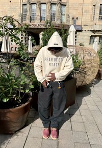 Creme hættejakke med "FEAR OF GOD STATE" trykt, sorte bukser og bordeaux støvler. Nøgleringe tilbehør synlige. Udendørs setting med grønne omgivelser.
