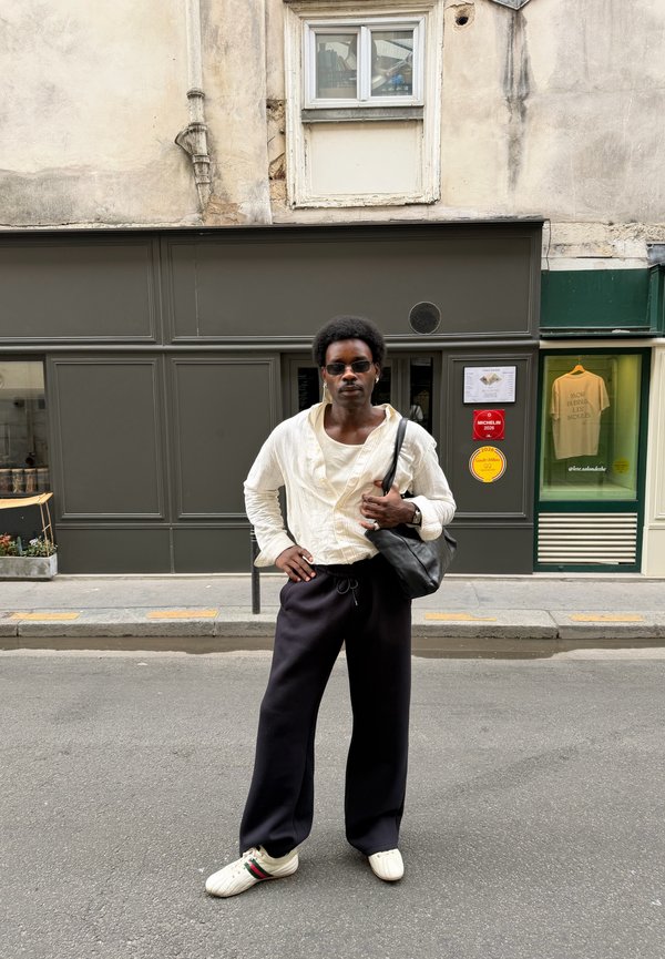 Persona con gafas de sol, camisa blanca, pantalones negros y zapatos blancos de pie en una calle frente a un edificio con fachada gris y ventana.