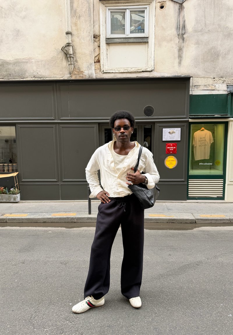 Persona con gafas de sol, camisa blanca, pantalones negros y zapatos blancos de pie en una calle frente a un edificio con fachada gris y ventana.