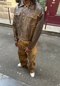 Άτομο με σκούρο καφέ δερμάτινο μπουφάν, καφέ φθαρμένες παντελόνες σε ευρεία γραμμή και λευκά παπούτσια, στέκεται σε έναν δρόμο με λιθόστρωτο.