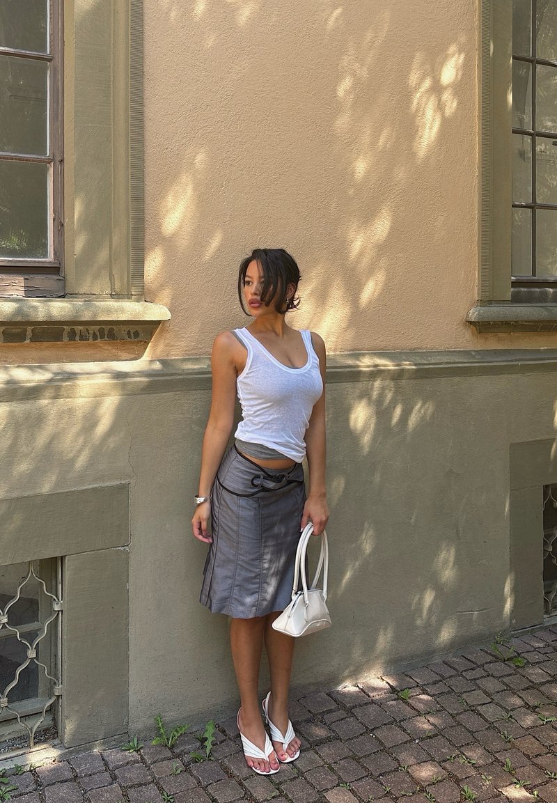 Frau in einem weißen Tanktop und einem grauen Rock, die eine weiße Handtasche hält und auf einem Kopfsteinpflaster neben einer beigen Wand mit Fenstern steht.