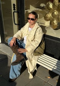 Bézs ballonkabát fehér póló felett, kék bő szárú farmernadrág, barna texturált térdig érő csizma. Zöld táska; napszemüveg, mintás körmök.