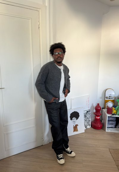 Hombre con gafas que lleva un cárdigan gris y pantalones negros está junto a una puerta blanca en una habitación con obras de arte y una pieza de ajedrez Supreme roja.
