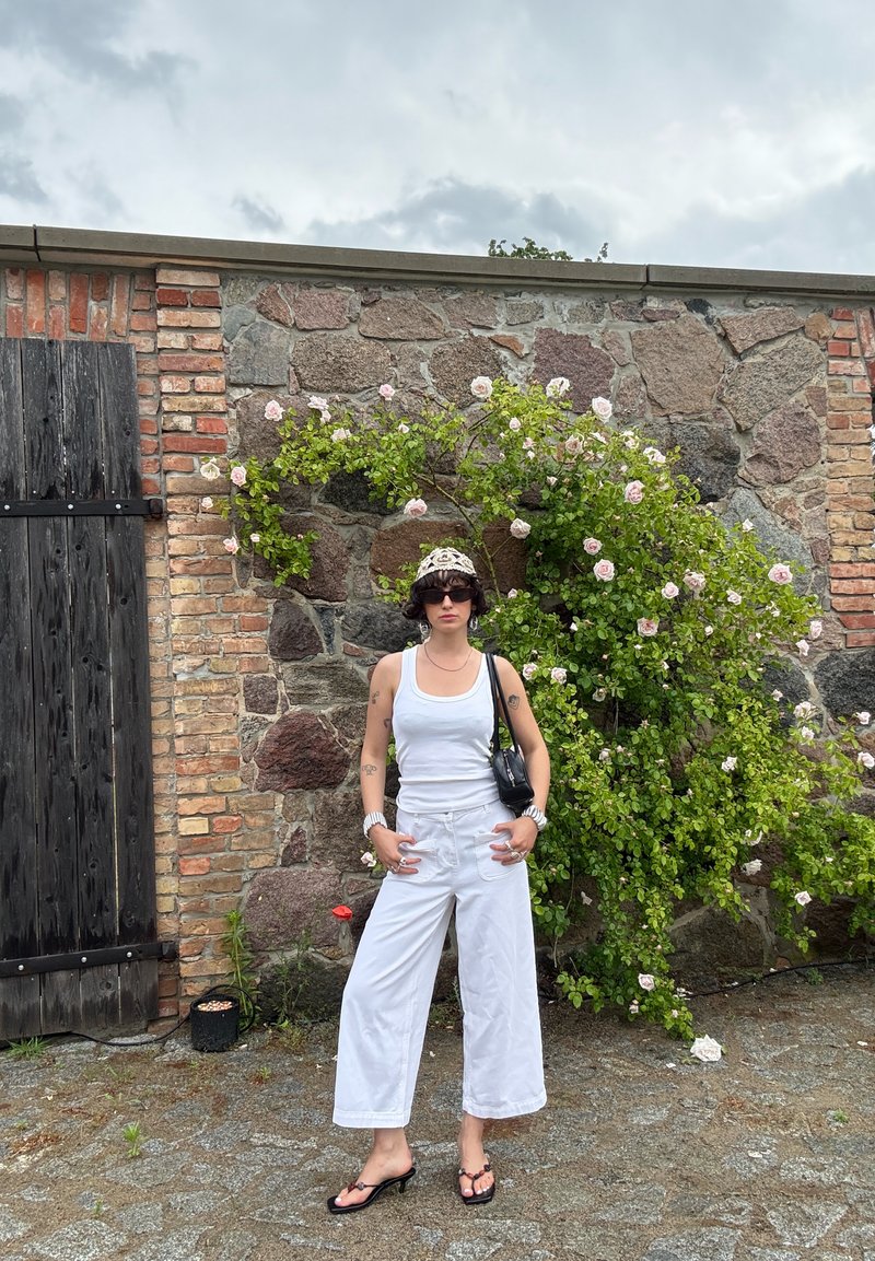 Personne en débardeur blanc et pantalon ample, sac à main noir et sandales, debout devant un mur en pierre avec des rosiers grimpants sous un ciel nuageux.