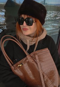Femme aux cheveux roux portant un chapeau en fourrure noire, de grandes lunettes de soleil noires, une écharpe beige et un manteau noir, tenant un sac à main marron texturé à l'intérieur près d'une fenêtre.