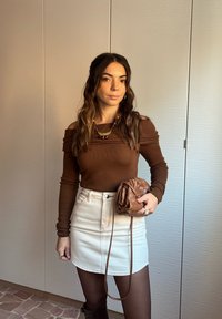 Hnědý top s odhalenými rameny, bílá džínová sukně a hnědá tkaná kabelka. Výrazné doplňky zahrnují vícebarevný náhrdelník a náušnice.