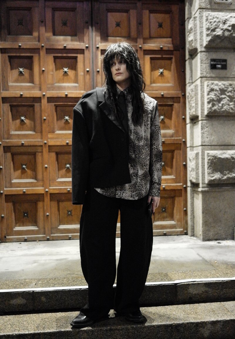 Veste noire oversize portée sur une chemise à motifs gris et blanc, associée à un pantalon large noir et des chaussures noires. Porte en bois avec des détails en forme d'étoile.