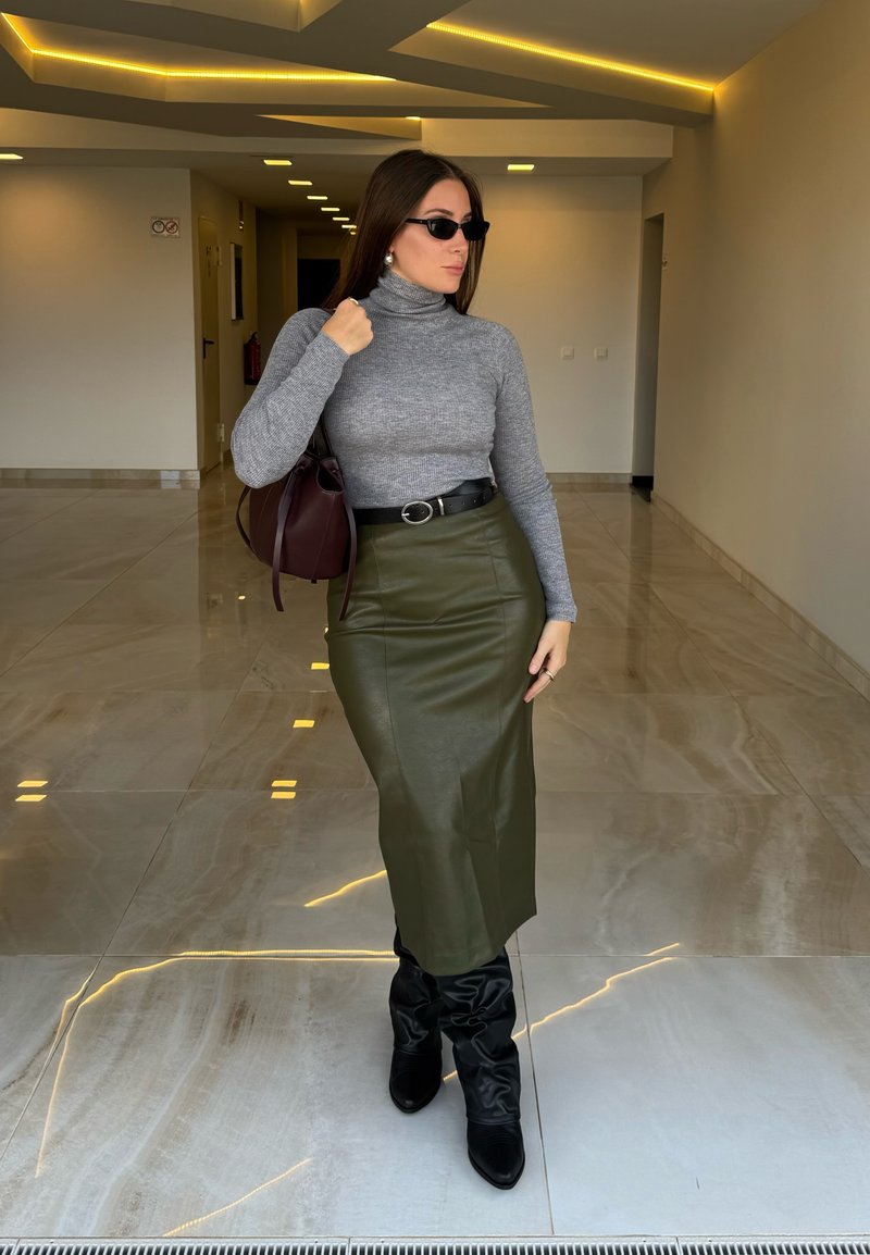 Donna in un maglione grigio attillato, gonna a matita in pelle verde oliva, stivali neri fino al ginocchio, che tiene una borsa a secchiello bordeaux, in una hall moderna.