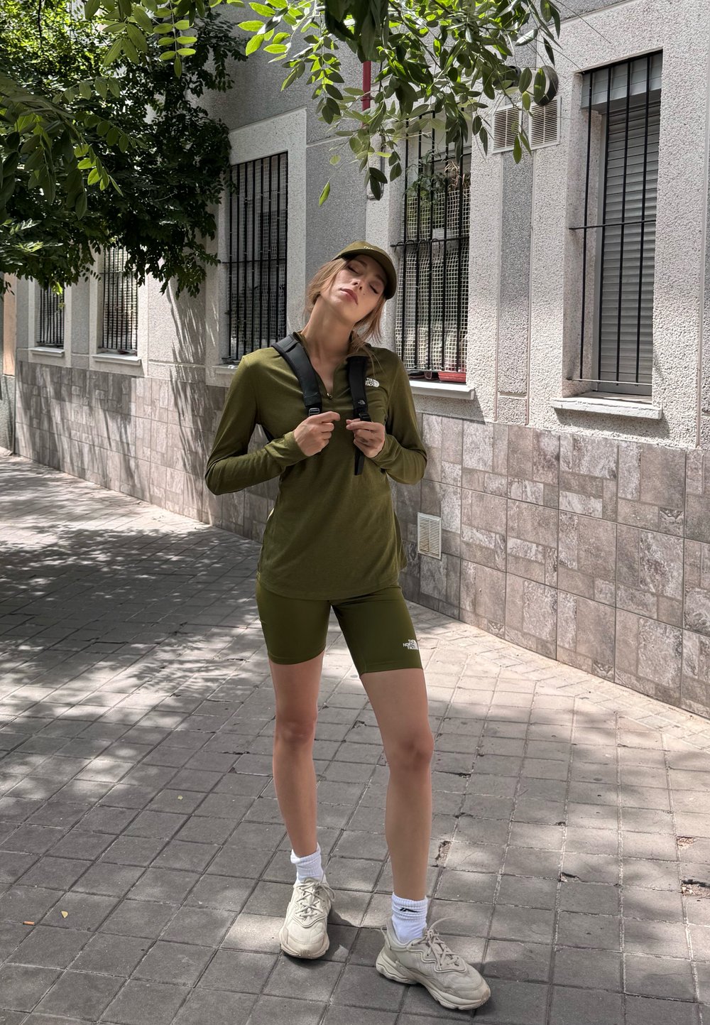 Conjunto deportivo en verde oliva con mangas largas y pantalones cortos de ciclista, acompañado de una gorra a juego. Zapatillas ligeras y calcetines blancos completan el look.