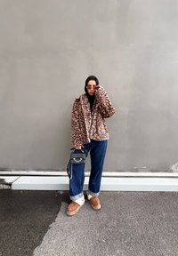 Ζακέτα από faux γούνα με λεοπάρ σχέδιο, μαύρο πουλόβερ, χαλαρά μπλε τζιν, καφέ slip-on παπούτσια, μαύρη τσάντα χειρός με αλυσίδα, γυαλιά ηλίου.