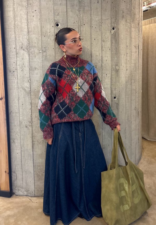 Mujer con un suéter de rombos colorido y una falda larga de denim sosteniendo una gran bolsa tote verde oliva junto a una pared de hormigón.
