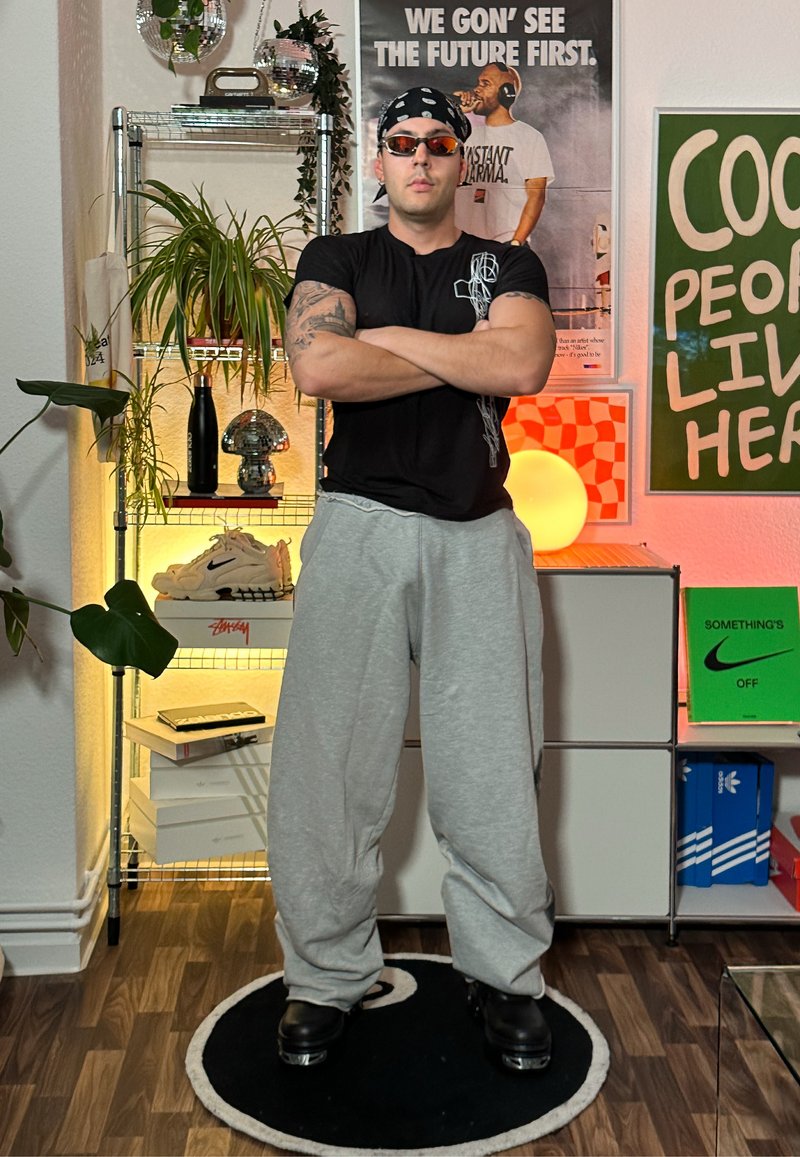 Black t-shirt, grey sweatpants, and black shoes. Accessories include a black bandana and sunglasses. Stands on a circular black rug.