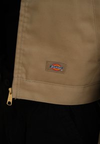 Beige tyg med en slät textur har en liten rektangulär Dickies-logotyp patch. En guldfärgad dragkedja finns längs kanten.