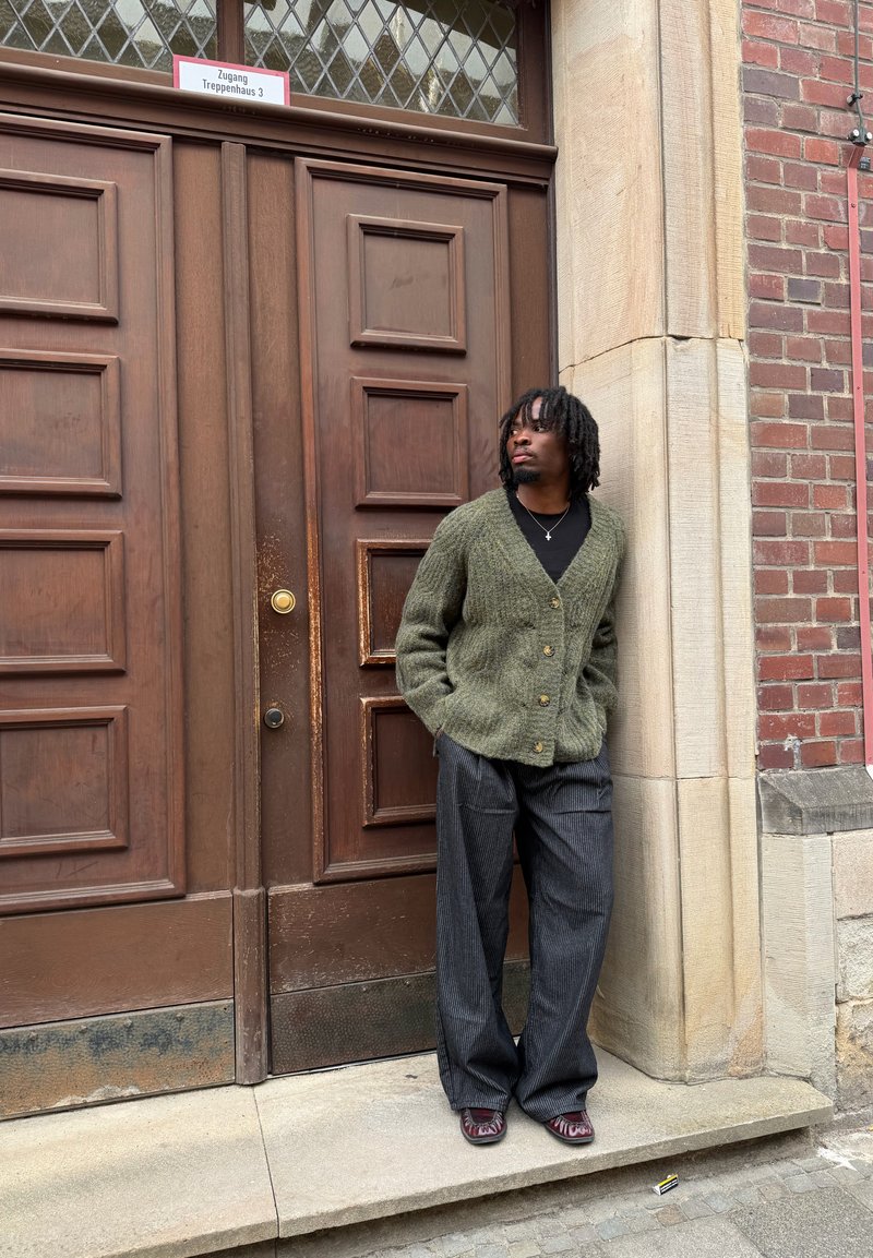 Homme avec des dreadlocks portant un cardigan vert et un pantalon large gris se tient contre un mur en pierre et en brique à côté d'une grande porte en bois avec une pancarte au-dessus.