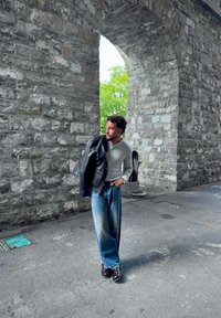Conjunto informal que incluye una camiseta henley gris, jeans de pierna ancha en azul deslavado y una chaqueta de cuero negra, ambientado en un entorno de arcos de piedra.