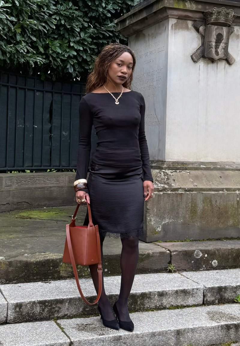 Frau in schwarzem Kleid und Strumpfhosen steht auf Steinstufen, hält eine braune Handtasche, im Hintergrund Grünpflanzen und ein steinernes Denkmal.