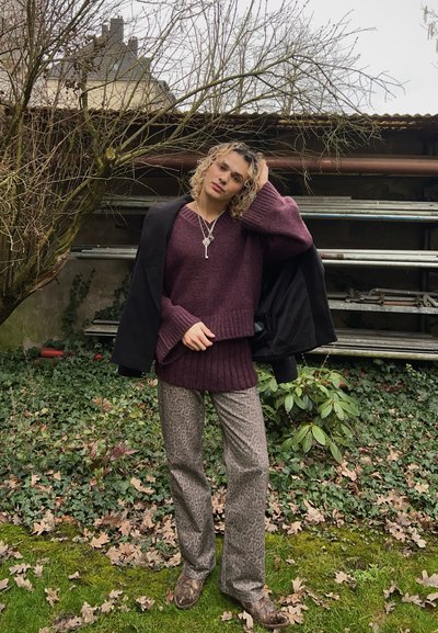 Suéter de punto burdeos con dobladillo acanalado combinado con pantalones de leopardo gris. Un abrigo negro colocado sobre el hombro, en un entorno natural al aire libre.