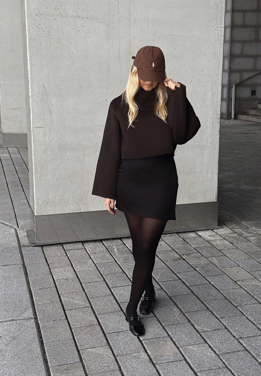 Svart kortärmad tröja med vida ärmar, kombinerad med en svart minikjol, svarta strumpbyxor och glänsande svarta skor. Brun keps med logov detalj.