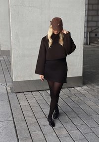 Svart kortärmad tröja med vida ärmar, kombinerad med en svart minikjol, svarta strumpbyxor och glänsande svarta skor. Brun keps med logov detalj.
