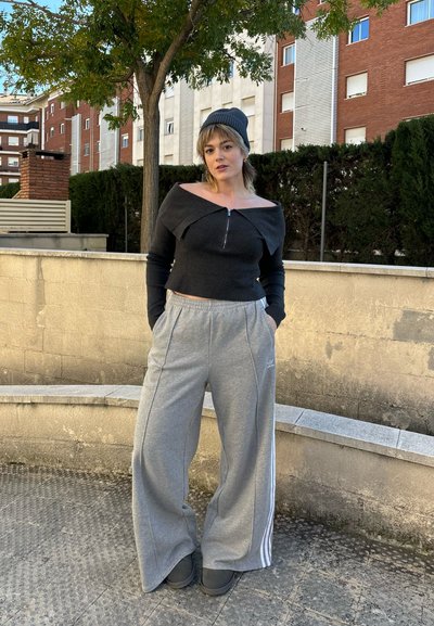 Parte superior negra de manga larga con diseño de hombros al descubierto y cremallera frontal, combinada con pantalones deportivos de pierna ancha grises claros con rayas verticales.