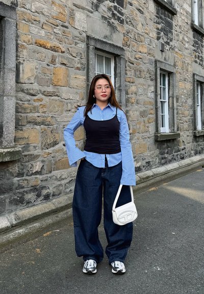 Joven mujer con gafas que lleva una camisa azul de rayas bajo un top negro, vaqueros anchos, zapatillas deportivas y sostiene un bolso blanco junto a una pared de piedra.