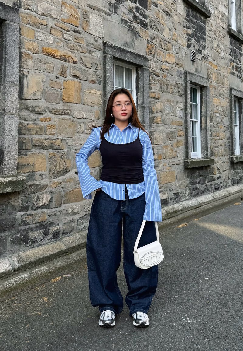 Junge Frau mit Brille trägt ein blau gestreiftes Hemd unter einem schwarzen Top, weite Jeans, Sneakers und hält eine weiße Handtasche an einer Steinmauer.
