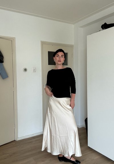 Mujer de pie en interiores vistiendo un top negro de manga larga, falda de satén blanca y zapatos negros, en una habitación minimalista con puertas cerradas.