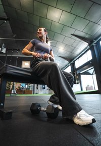 Svart heksagonal manual på gulvet i treningsstudioet ved siden av en person som sitter på en svart benk, iført grå topp og løse grå bukser med hvite joggesko.