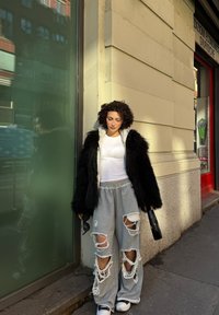 Svart oversized jakke i falsk pels, hvit tettsittende topp, grå sweatpants med revner, hvite joggesko, holder en veske, står foran en glassvegg.