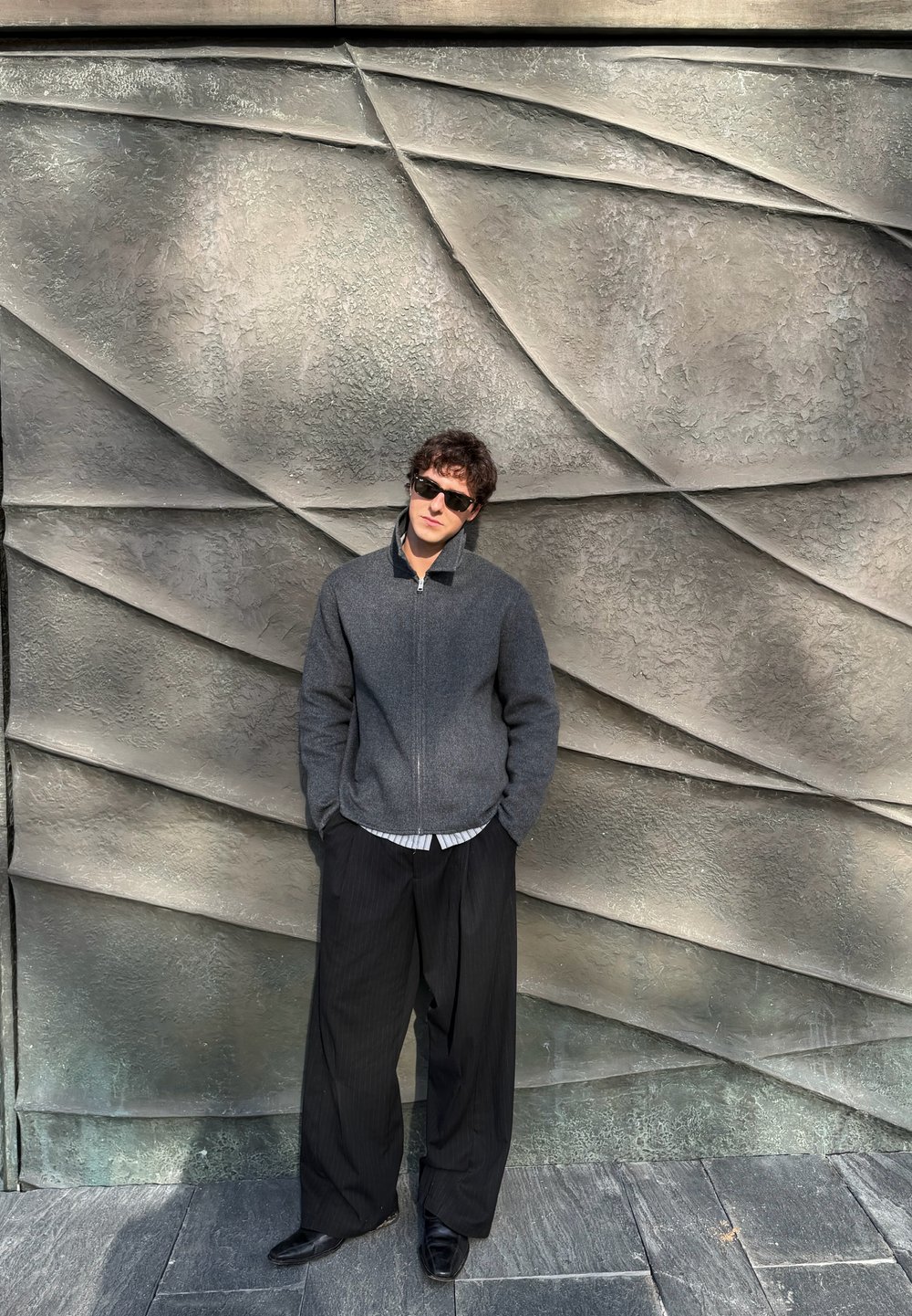 Jeune homme portant des lunettes de soleil, une veste grise zippée et un pantalon large noir, debout les mains dans les poches devant un mur en pierre texturé aux formes géométriques.